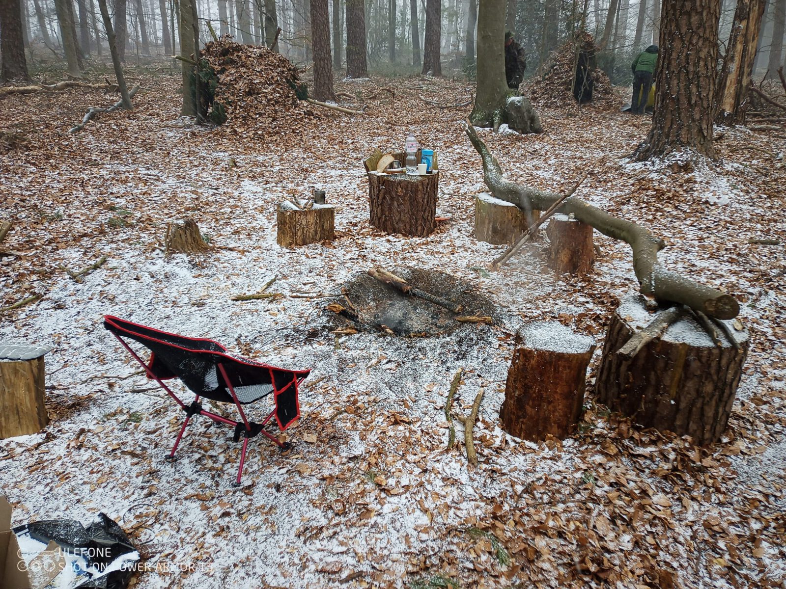 alt=“Lagerstelle bei einem Survivalkurs mit erloschener Feuerstelle und Notunterkünften im winterlichen Wald″