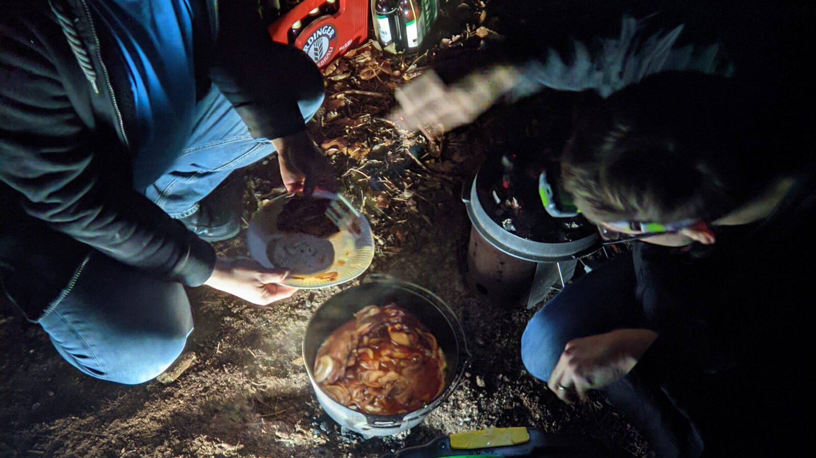 alt=“Zwei Menschen sitzen in dunklem Wald nehmen sich essen aus dem Dutchoven″