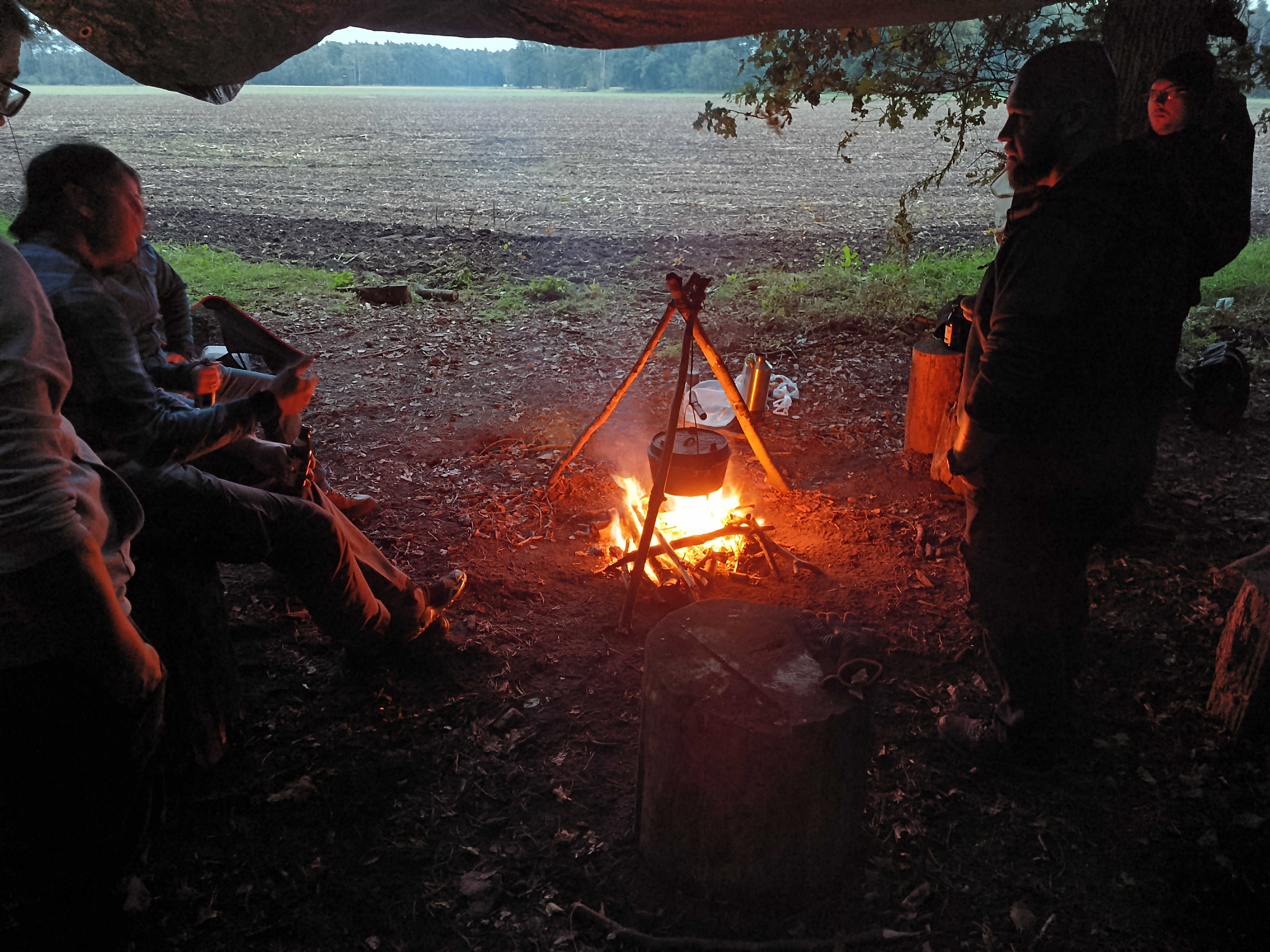 alt="Lagerplatz mit Lagerfeuer am frühen Abend. Über dem Feuer hängt ein Topf im Hintergrund sieht man ein Feld."